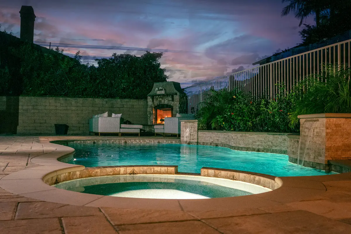 A lit private outdoor pool at dusk, with a fire feature and terrace beyond the water