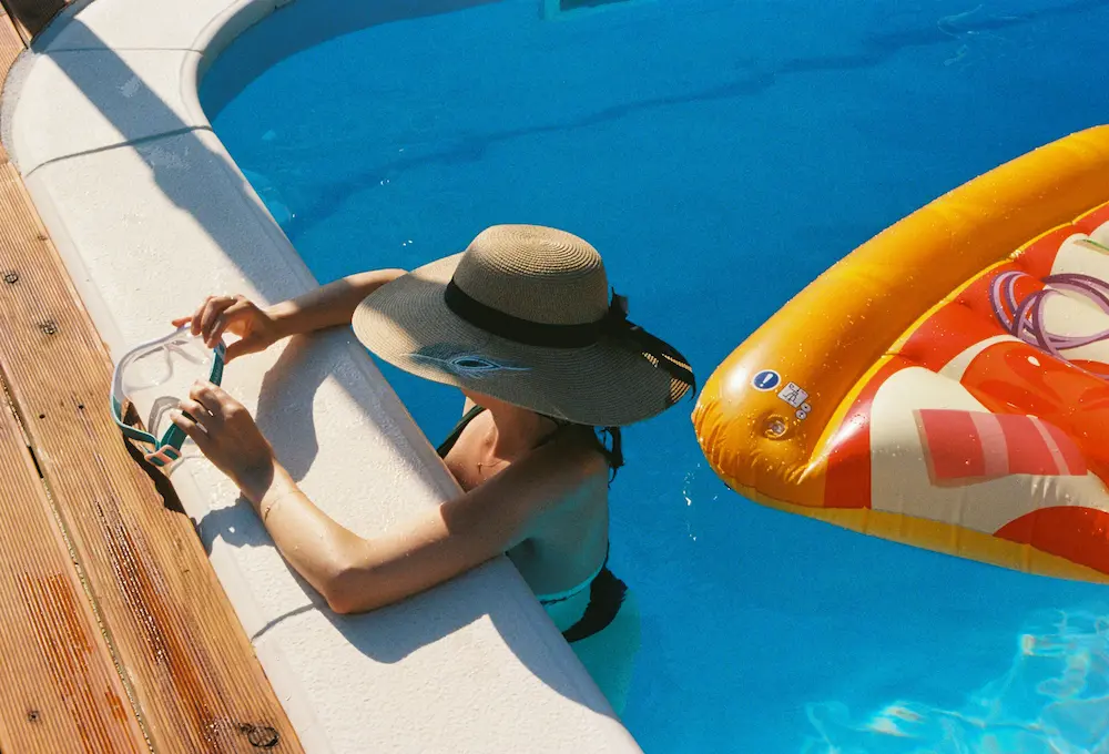 The edge of a private pool on a summer afternoon — a swimmer in a sun hat rests at the wall beside an inflatable